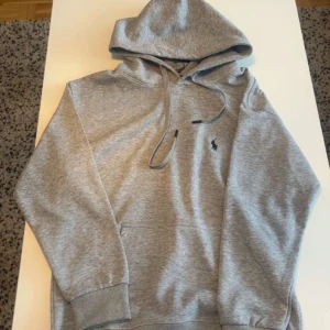 Grå  Polo Ralph Lauren Hoodie - Säljer en klassisk grå hoodie från Polo Ralph Lauren med broderad logga på bröstet. Tröjan har huva med dragsko och ribbade muddar vid ärmslut och nederkant. Perfekt för en avslappnad stil.