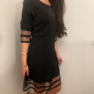 Svart klänning med transparenta detaljer - Säljer en svart klänning med halvtransparenta ränder på ärmar och nederkant. Klänningen har rund halsringning och trekvartsärm. Materialet känns slätt och lätt, perfekt för dig som gillar stilrena plagg med en twist. Den har även en lång dragkedja under arman som ger dig fina former
