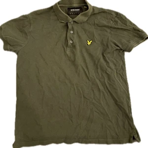 Olivgrön pikétröja från Lyle & Scott - Säljer en olivgrön pikétröja från Lyle & Scott med klassisk krage och tre knappar framtill. Tröjan har korta ärmar och den ikoniska gula loggan broderad på bröstet. Perfekt för en avslappnad och stilren look.