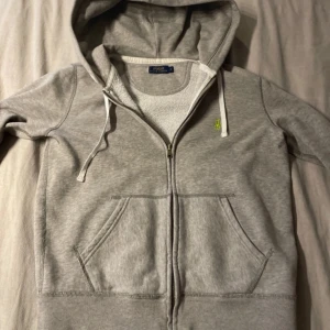 Grå hoodie från Polo Ralph Lauren - Säljer en klassisk grå hoodie från Polo Ralph Lauren med dragkedja och huva. Tröjan har fickor framtill och en broderad grön logga på bröstet. Perfekt för en avslappnad stil.