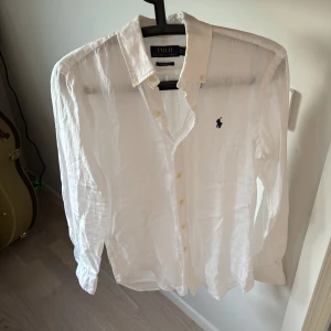 Vit linneskjorta från Polo Ralph Lauren - Klassisk vit skjorta i linne från Polo Ralph Lauren  med broderad marinblå logga på bröstet. Skjortan har lång ärm, knappar framtill och klassisk krage. Perfekt för en stilren och fräsch look. 