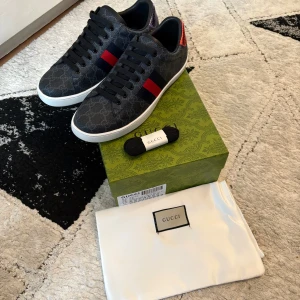 Gucci ace skor  - Säljer ett par svarta Gucci sneakers med diskret GG-mönster, röd och blå rand på sidan samt röd detalj vid hälen. Skorna har rund tå, platt vit sula och kommer med extra svarta skosnören och dustbag.Priset kan självklart diskuteras och de är sprillans nya 