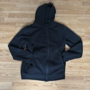Svart hoodie med dragkedja från Dressmann - Säljer en svart hoodie från Dressmann i storlek S. Jackan har dragkedja framtill, huva med snörning och ribbade muddar. Perfekt för en avslappnad stil och enkel att matcha med det mesta.