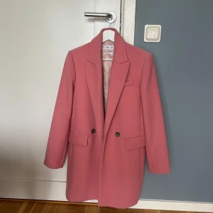Rosa kappa från Mango - Säljer en snygg rosa kappa från Mango med klassisk krage och dubbelknäppning framtill. Kappan har långa ärmar, två fickor och är fodrad. Perfekt för dig som vill ha en stilren och trendig look. Helt oanvänd i bra material / ull.