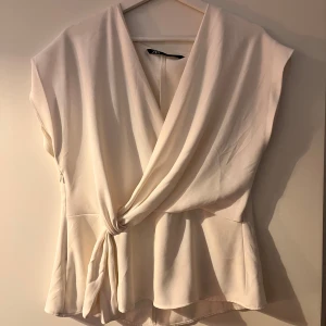 Topp från Zara - Säljer en krämvit omlottopp från Zara med knyt i midjan och lös passform. Toppen har korta ärmar och ett snyggt fall. Perfekt för vår och sommar, enkel att styla till olika looks.
