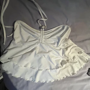 Vit volang topp med halterneck - Supersöt vit topp med halterneck och volanger i flera lager. Toppen har en snygg rynkning framtill och knytband i nacken. Perfekt för sommarens alla tillfällen när du vill känna dig extra fin. 