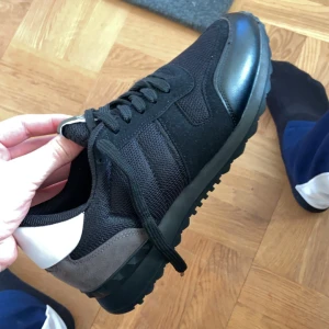 Svarta sneakers med vita och grå detaljer - Säljer ett par snygga svarta Valentino sneakers med vita och grå detaljer. Skorna har svarta snören, rund tå och platt sula med mönstrad undersida. Perfekta för dig som vill ha en stilren och sportig look. Priset kan diskuteras vid snabb affär