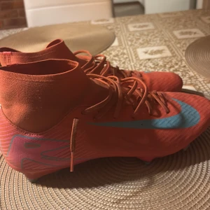 Röda Nike fotbollsskor med blå swoosh - Säljer ett par röda Nike fotbollsskor med blå swoosh och högt skaft. Skorna har snörning och är designade för bra grepp på planen. Perfekta för dig som vill sticka ut på fotbollsplanen!