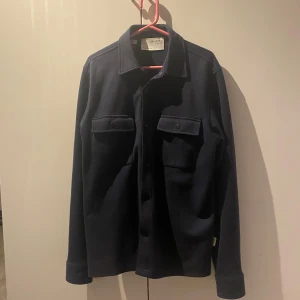 Mörkblå overshirt från Selected Homme - Säljer en stilren mörkblå overshirt från Selected Homme. Skjortan har två bröstfickor med lock och knappar samt klassisk krage. Perfekt att bära som lager över en t-shirt eller tröja.
