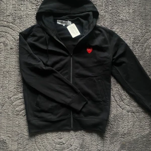 Comme des Garçons Play zip-uo - Svart hoodie från Comme des Garçons Play med dragkedja och det ikoniska röda hjärtat broderat på bröstet. Tillverkad i 100% bomull och har långa ärmar samt huva med dragsko. Perfekt för en stilren och avslappnad look. 🎶HELT NY!!🎶