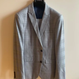 Grå rutig kostym från Lindbergh - Snygg grå kostym med rutigt mönster från Lindbergh White. Kavajen har två knappar framtill, klassisk krage och fickor med lock. Byxorna har pressveck och knappdetalj bak. Perfekt för bal eller student.