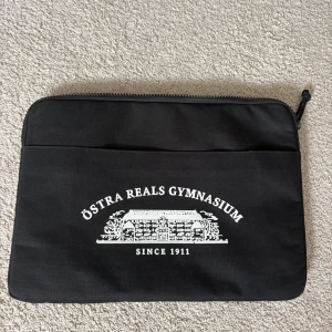 Svart datorväska Östra Reals Gymnasium - Svart datorväska med vit tryckt logga och texten 'Östra Reals Gymnasium Since 1911' på framsidan. Väskan har dragkedja upptill och ett extra fack på framsidan. Perfekt för att bära med sig laptop och skolmaterial.