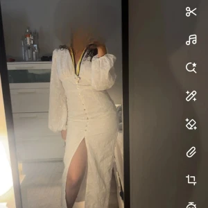 Vit långklänning med slits och puffärm - Supersnygg vit långklänning med v-ringning, puffiga långärmar och markerad midja. Klänningen har knappar framtill och en hög slits som ger en extra touch. Broderade blommor i tyget och romantisk känsla. Perfekt för dig som vill sticka ut!