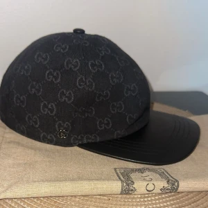 Gucci Dubai Noir keps - Snygg svart keps från Gucci med klassiskt GG-monogram över hela ovandelen. Skärmen är i svart skinn och kepsen har justerbart spänne bak. Materialmix av bomull och skinn ger en lyxig känsla. Perfekt accessoar för att lyfta din outfit. Dustbag medkommer, fler bilder privat! Öppen via byten (Göteborg).