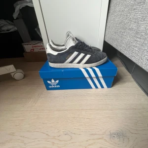 Adidas Gazelle sneakers marinblå/vit - Säljer ett par klassiska Adidas Gazelle sneakers i marinblå mocka med vita detaljer och de ikoniska tre ränderna på sidan. Skorna har snörning och platt sula, samt diskret guldfärgad Gazelle-text på sidan. Perfekta för en stilren och sportig look.