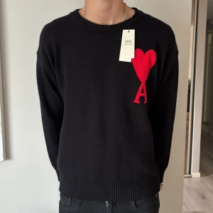 Svart stickad tröja från Ami Paris - Stickad tröja i nytt skick från Ami Paris med röd hjärtlogga och bokstaven A på bröstet. Klassisk rund halsringning, ribbade muddar och lång ärm. Perfekt för dig som gillar stilrena plagg med en twist. Materialet är mjukt och skönt mot huden.