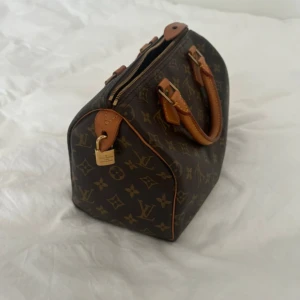 Louis Vuitton speedy 25 väska  - Säljer en klassisk Louis Vuitton handväska i brunt skinn med ikoniskt monogrammönster och detaljer i ljusbrunt läder. Väskan har två handtag, guldfärgad dragkedja och hänglås. Perfekt storlek för det viktigaste och en riktig statement piece. Den är i storlek 25 och bra skick. Tillverkad år 1995. Hänglås och orginal nyckel finns. 