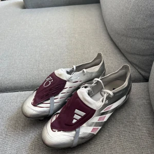 Adidas Predator fotbollsskor silver/vinröd - Säljer ett par Adidas Predator fotbollsskor i storlek 44. Skorna är silverfärgade med vinröda detaljer och har snörning samt täckflik med Predator-logga. Kommer med tillhörande vinröd påse. Perfekta för dig som vill sticka ut på planen! Använda ett fåtal gånger. Säljer pågrund av att de är för små. Köpt på unisport för ca 2800kr