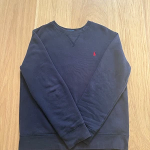 Mörkblå sweatshirt från Polo Ralph Lauren - Klassisk mörkblå sweatshirt från Polo Ralph Lauren med rund halsringning och röd broderad logga på bröstet. Tröjan har långa ärmar och är tillverkad i mjuk bomull, perfekt för en avslappnad och stilren look. allt superb utan en ytterst litet hål vid ryggen 
