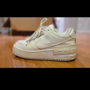 Nike Air Force 1 Shadow vita sneakers - Nike Air Force 1 Shadow i helvitt skinn med chunky plattformssula och klassisk swoosh. Snygga detaljer som dubbla lager och präglad AIR-text på hälen. Perforerad tå och vita skosnören ger en clean look. Perfekta för dig som gillar streetstyle.