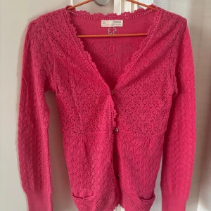 Rosa kofta från Odd Molly - Supersöt rosa kofta från Odd Molly med virkade spetsdetaljer, knappar framtill och knytband i ryggen. Långärmad och stickad i bomull, med dekorativa mönster och små fickor framtill. Perfekt för dig som gillar unika och färgglada plagg!