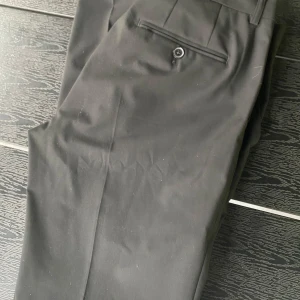 Svarta kostymbyxor Dressmann 32/32 - Mycket fina svarta kostymbyxor från Dressmann i slim fit-modell. Klassisk design med pressveck, bälteshällor och bakficka med knapp. Använda två gånger.