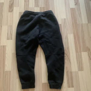 Svarta mjukisbyxor från Nike - Svarta mjukisbyxor från Nike med resår i midjan och muddar vid bensluten. Byxorna har en enkel design med diskret Nike-logga på ena benet och dragkedjeförsedda fickor. Perfekta för chill eller träning.