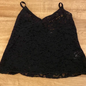 Svart spetslinne från Kappahl - Svart linnetopp från Kappahl i storlek M. Toppen har tunna axelband och är helt täckt av vacker spets med blommigt mönster. Perfekt att styla med jeans eller kjol för en snygg look.