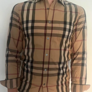 Rutig Burberry skjorta i bomull - Säljer en klassisk rutig skjorta från Burberry i beige, svart, vit och röd. Skjortan har lång ärm, knappar framtill och bröstficka. Den är tillverkad i mjuk bomull och har den ikoniska Burberry-rutan. Skick 9/10, använder ej då den har blivit för liten. Storlek S