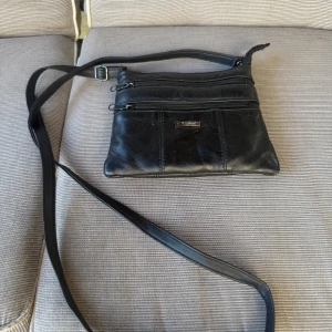 Svart axelväska från Feel Accessories - Svart axelväska i skinn från Feel Accessories med två dragkedjefack framtill och justerbar axelrem. Stilren design med metallmärke på framsidan. Perfekt storlek för mobil, plånbok och nycklar.
