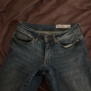 Blåa ultra low bootcut jeans från Vero Moda - Säljer ett par snygga blå jeans från Vero Moda med klassisk femficksdesign och bootcut-ben. Jeansen har en mörk tvätt med lätt slitningar och är tillverkade i jeansmaterial med dragkedja och knapp framtill. Perfekt för dig som gillar retrovibbar och vill ha ett coolt statement.