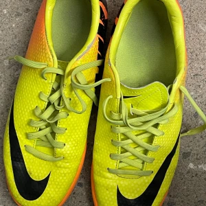Nike Mercurial fotbollsskor gul/orange - Säljer ett par Nike Mercurial fotbollsskor i storlek 39. Skorna är i en riktigt cool gul färg med orange detaljer och svarta Nike-loggor. De har snörning och är tillverkade i syntetmaterial, perfekta för dig som vill sticka ut på planen.