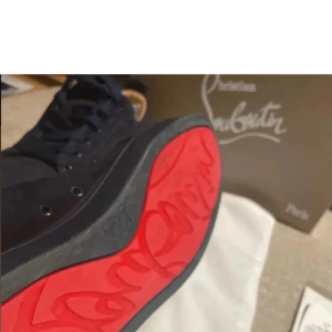 Christian Louboutin svarta sneakers mocka - Exklusiva svarta sneakers från Christian Louboutin i mjuk mocka med ikonisk röd sula och broderad logga på hälen. Snygg och stilren design med rund tå och snörning. Perfekta för dig som vill sticka ut med lyxiga detaljer.