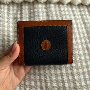 Dooney & Bourke plånbok - Äkta plånbok från Dooney & Bourke från 90-talet i marinblå och brunt skinn med klassisk logga framtill. Plånboken har en stilren design med bruna kanter, fack för kort och mynt samt ett transparent fönster för ID. Perfekt storlek för fickan och riktigt tidlös vibe. 😍