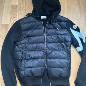 Moncler cardigan  - Snygg svart cardigan från Moncler med stickade ärmar och huva. Jackan har dragkedja framtill, coolt blixtmönster i grått på ena ärmen och Moncler-logga. Perfekt för dig som vill ha en streetig och exklusiv look. Inga tecken på användning.                                                                  !PRIS KAN SÄNKAS VID SNABB AFFÄR!