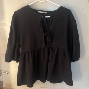 Svart blus  - Svart blus från  med trekvartsärmar och knytning framtill. Blusen har en lös passform, volang nedtill och är tillverkad i ett lätt, strukturerat tyg som ger en avslappnad vibe.