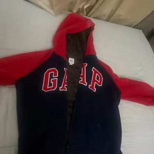 Blå och röd hoodie från GAP Kids - Snygg hoodie från GAP Kids i marinblått med röda ärmar och huva. Stor GAP-logga framtill, dragkedja och mjukt, grått fleecefoder på insidan. Perfekt för kyliga dagar och riktigt skön att ha på sig. Klassisk och sportig stil med känguruficka.