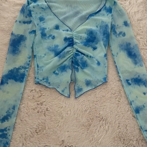 Blå tie-dye mesh topp  - Säljer en blå mesh topp med lång ärm och snygg tie-dye effekt i olika blå nyanser. Toppen har v-ringning och cool snörningsdetalj framtill som ger en rynkad look. Perfekt för dig som gillar unika och trendiga plagg!