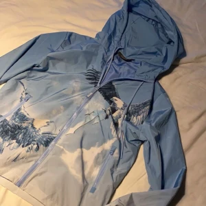 Trapstar vindjacka - Snygg blå vindjacka från Trapstar med stort örntryck och broderad logga på ryggen. Jackan har huva, dragkedja framtill och coolt motiv i vitt och blått som ger streetkänsla. Bara att höra av er vid frågor. 