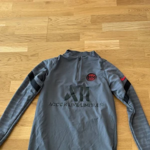 PSG tröja - Snygg grå långärmad träningströja från Nike och Paris Saint-Germain med svart tryck, röd PSG-logga och diskret mönster på ärmarna. Half zip framtill och svart Nike-logga på ärmen. Perfekt för fotbollsträning eller chill. Storlek S, hör av er vid fler frågor :)