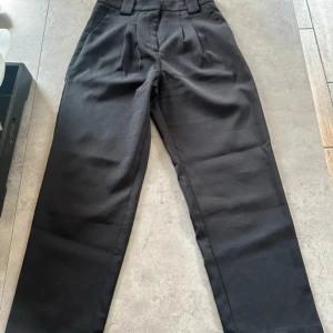 Svarta kostymbyxor från NA-KD - Snygga svarta kostymbyxor från NA-KD med hög midja och raka ben. Byxorna har pressveck framtill och breda hällor i midjan. Perfekta för en clean och stilren look. Materialet känns mjukt och följsamt.