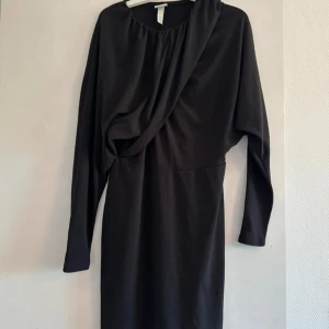 Svart långärmad klänning H&M S - Svart långärmad klänning från H&M i storlek S. Klänningen har en draperad omlott-detalj framtill och är figurnära med markerad midja. Mjuk och stretchig känsla, perfekt för en stilren look.