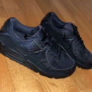 Svarta Nike Air Max 90 sneakers - Säljer ett par helsvarta Nike Air Max 90 sneakers med klassisk chunky sula och synlig Air-enhet. Skorna har ovandel i mesh och skinn, med svarta snören och diskreta detaljer. Perfekta för dig som gillar en clean och sportig look.