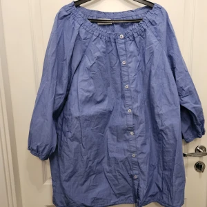 Bomullsblus från Ulla Popken i storlek 50/52 - Blå blus i bomull från Ulla Popken med trekvartsärm och knappar framtill. Den har en rynkad halsringning och dragsko nedtill. Mycket gott skick. Storlek 50/52.  Bröstvidd 120 cm Längd 75 cm