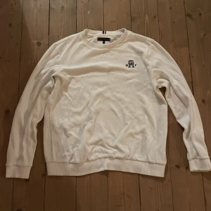 Vit tröja Tommy Hilfiger 164 - Vit långärmad tröja från Tommy Hilfiger med broderad logga på bröstet och diskret flagga på ärmslutet. Tröjan har ribbade muddar och krage samt en klassisk, stilren look. Materialet är mjukt och känns skönt mot huden.