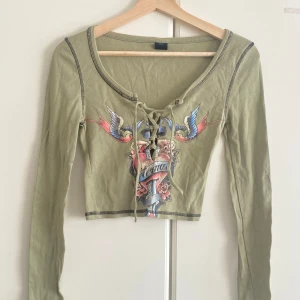 Olivgrön croppad topp Urban Outfitters - Olivgrön croppad långärmad topp från Urban Outfitters med coolt tatueringsinspirerat tryck framtill, snörning vid urringningen och kontrastsömmar. Perfekt för dig som gillar edgy och unik stil.