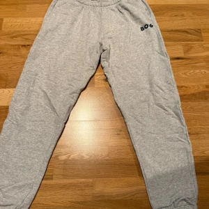 Grå mjukisbyxor med BOS-tryck - Säljer ett par grå mjukisbyxor med elastisk midja och svart BOS-tryck på ena höften. Byxorna är i mjukt material och har en avslappnad passform, perfekta för chill dagar eller träning.