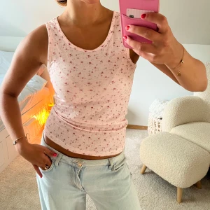 Ljusrosa blommig linnetopp, från NOELLA - Supersöt ljusrosa linnetopp med små blommor i rosa och rött. Toppen är ribbad och har en tight passform med ärmlös design och rund halsringning. Perfekt för varma dagar och enkel att matcha med jeans eller kjol.
