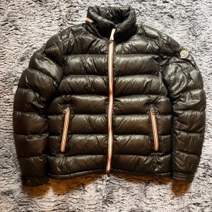 Svart Moncler pufferjacka herr - Snygg svart pufferjacka från Moncler med klassisk quiltad design och hög krage. Jackan har dragkedja med röd-vita detaljer, två fickor med dragkedja och Moncler-logga på ärmen. Perfekt för kalla dagar och riktigt stilren vibe.