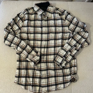 Rutig skjorta från Dressmann M - Snygg rutig skjorta från Dressmann i storlek M. Skjortan har ett klassiskt rutmönster i beige, svart och brunt med vita inslag. Långärmad modell med knappar framtill och två bröstfickor. Perfekt för en avslappnad men ändå stilren look.